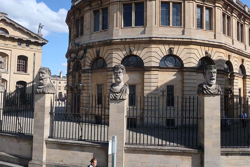 The heads sit on the wall and fence that surrounds the Sheldonian Theatre.